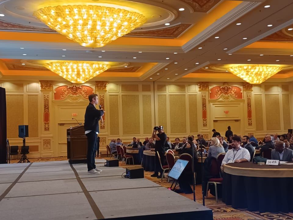 Joe Cummings hosting a ballroom conference on stage with a microphone before a large seated audience