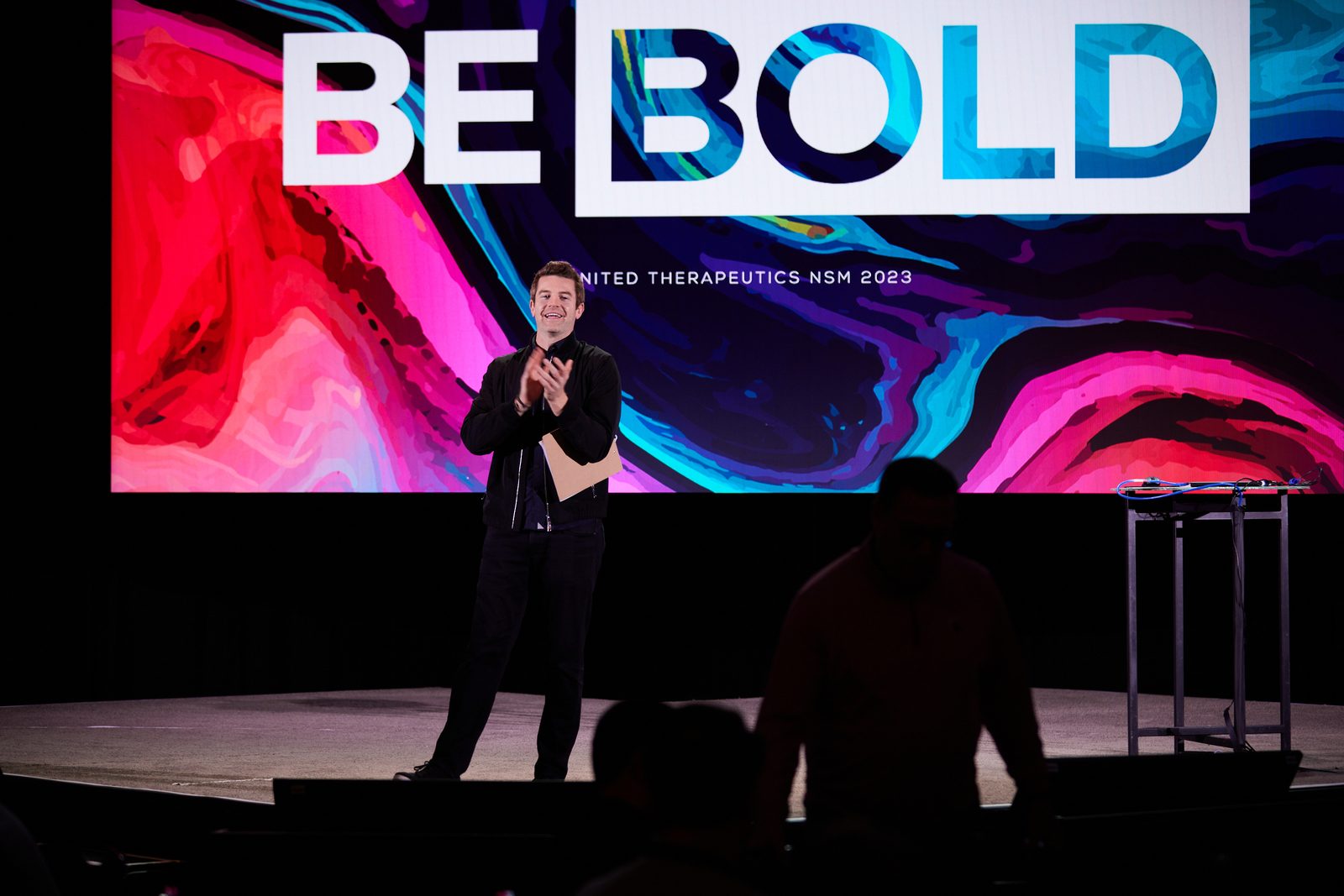 Joe Cummings presenting on a large corporate conference stage in front of a BE BOLD branded screen