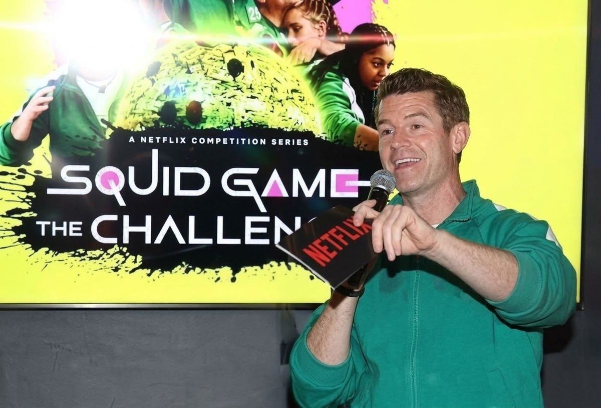 Joe Cummings in a green tracksuit hosting a Netflix Squid Game The Challenge event with a microphone