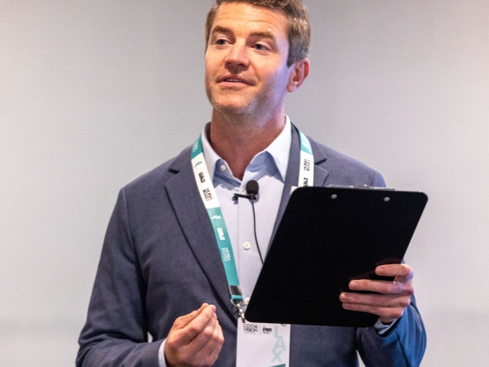 Joe Cummings with a clipboard and conference lanyard moderating a panel at a 2024 vision conference
